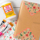 A kraft paper notebook or journal with floral washi tape borders in coral, pink, green, and cream, shown on a white wood background with a bottle of Mod Podge and two paintbrushes.