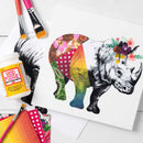 A rhinoceros art print featuring a black-and-white rhino with a colorful patchwork body and floral crown, styled on a white surface with Mod Podge, paintbrushes, and paint swatches.